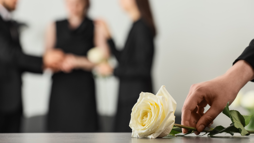 Dłoń kładąca białą różę na trumnie podczas ceremonii pogrzebowej.